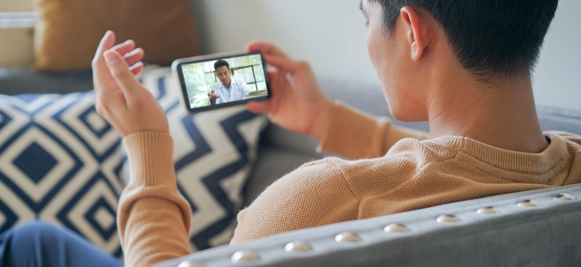 May show: A person sits on a couch holding a smartphone and watching a video call or online video featuring another individual.
