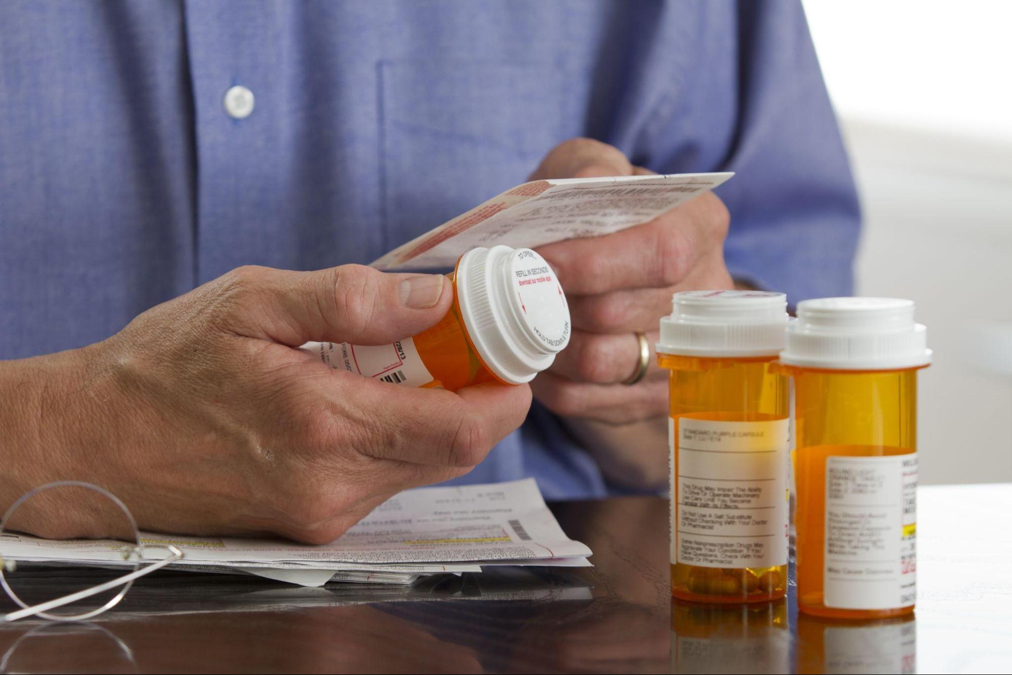 May show: A person in a blue shirt holds a prescription bottle and reads medication instructions at a table with several other prescription bottles.
