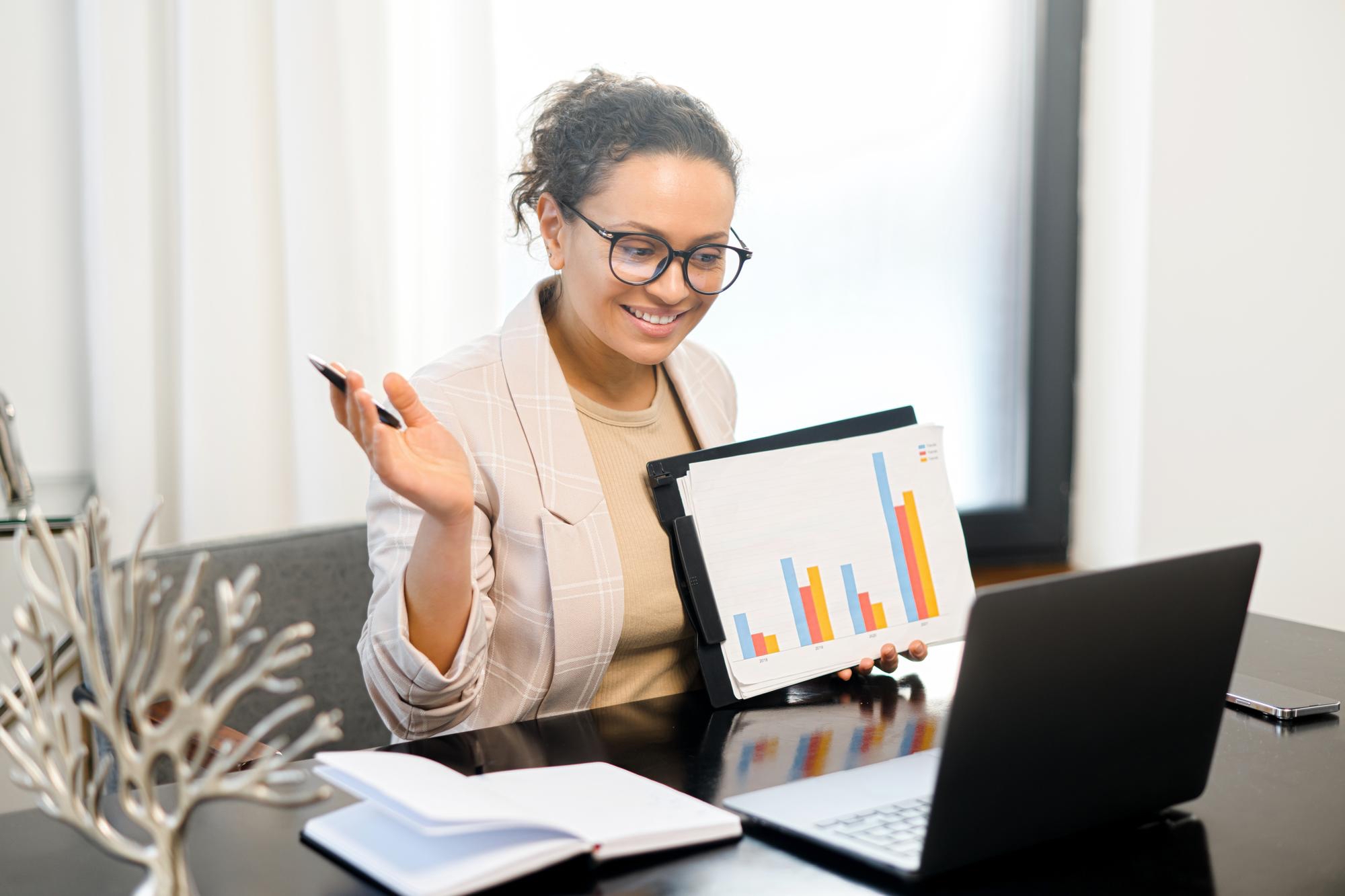 A woman wearing glasses sits at a desk, smiling at her laptop while holding up a sheet with colorful bar graphs during a video call.