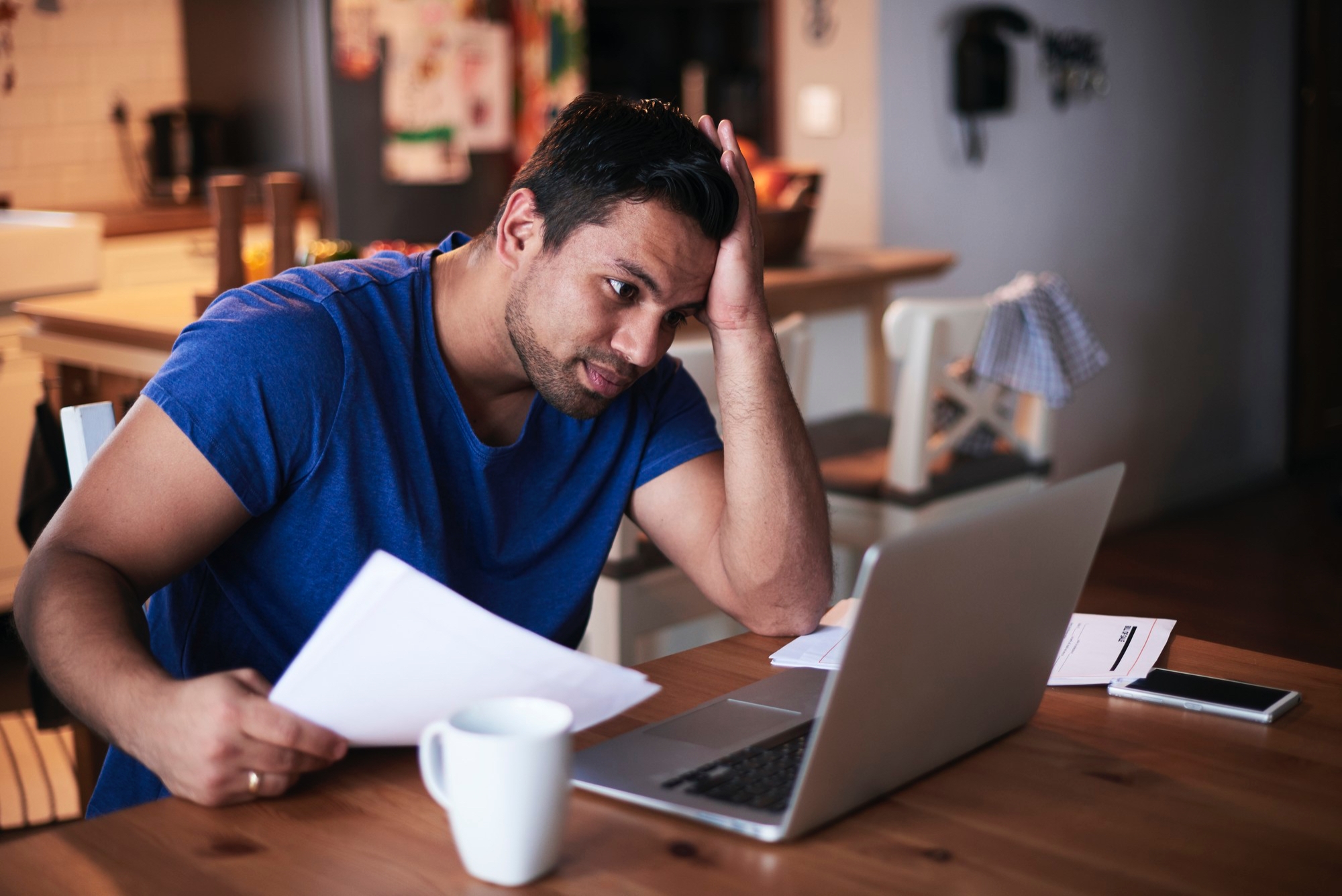 A man sits at a table holding papers and looking at a laptop with a concerned expression. A coffee mug, documents, and phone are on the table.