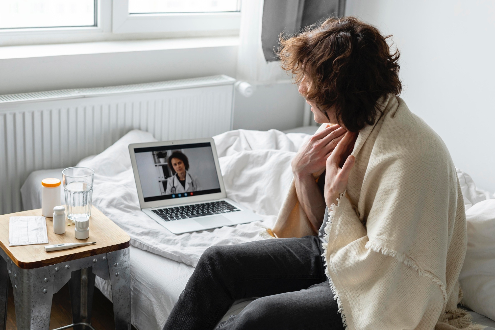A person wrapped in a blanket sits on a bed having a video call with a doctor, holding their throat. Medicine, a glass of water, and tissues are on a table nearby.