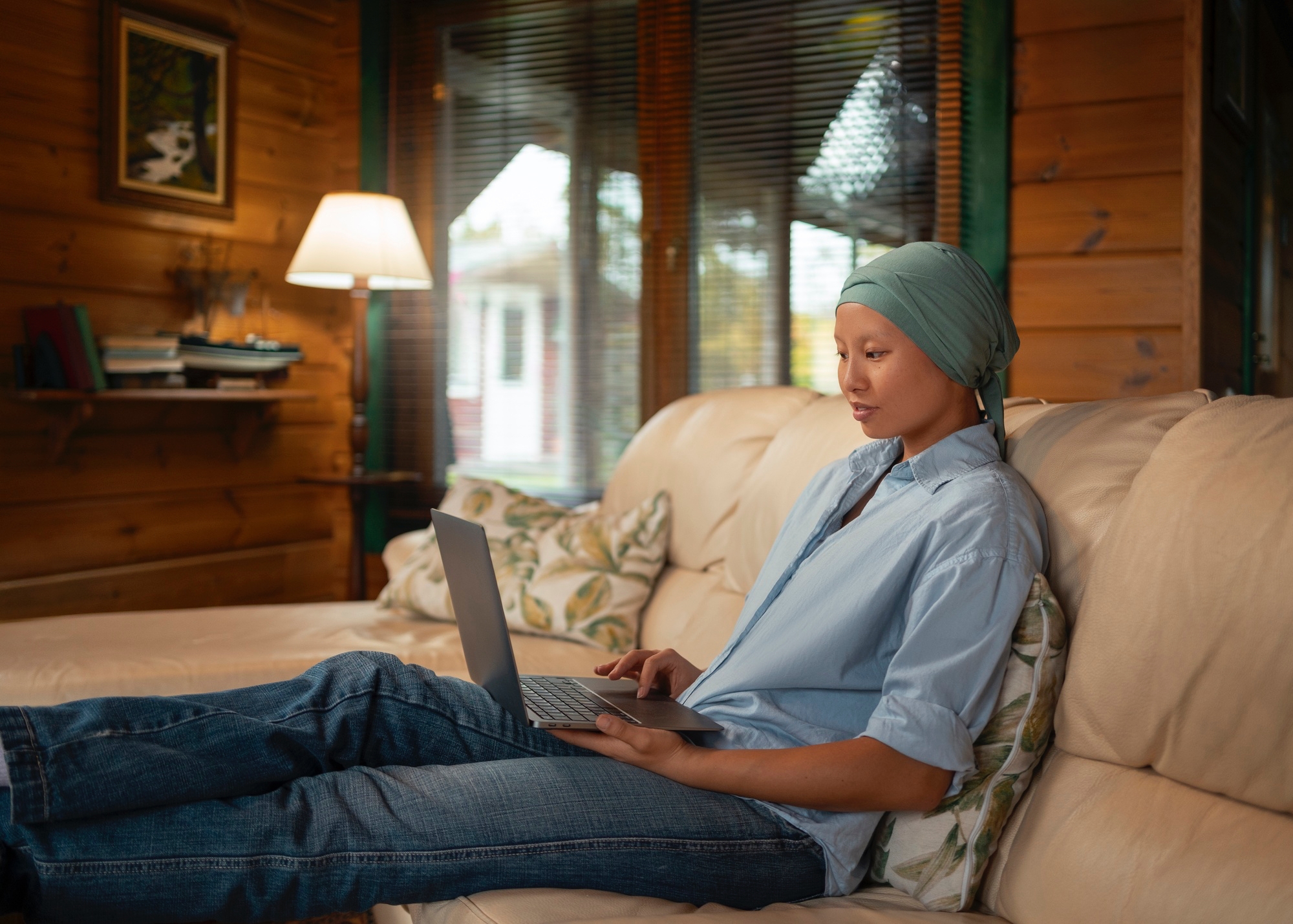 A woman sits on a sofa in a wood-walled room, wearing a headscarf and using a laptop, with a window and lamp in the background.