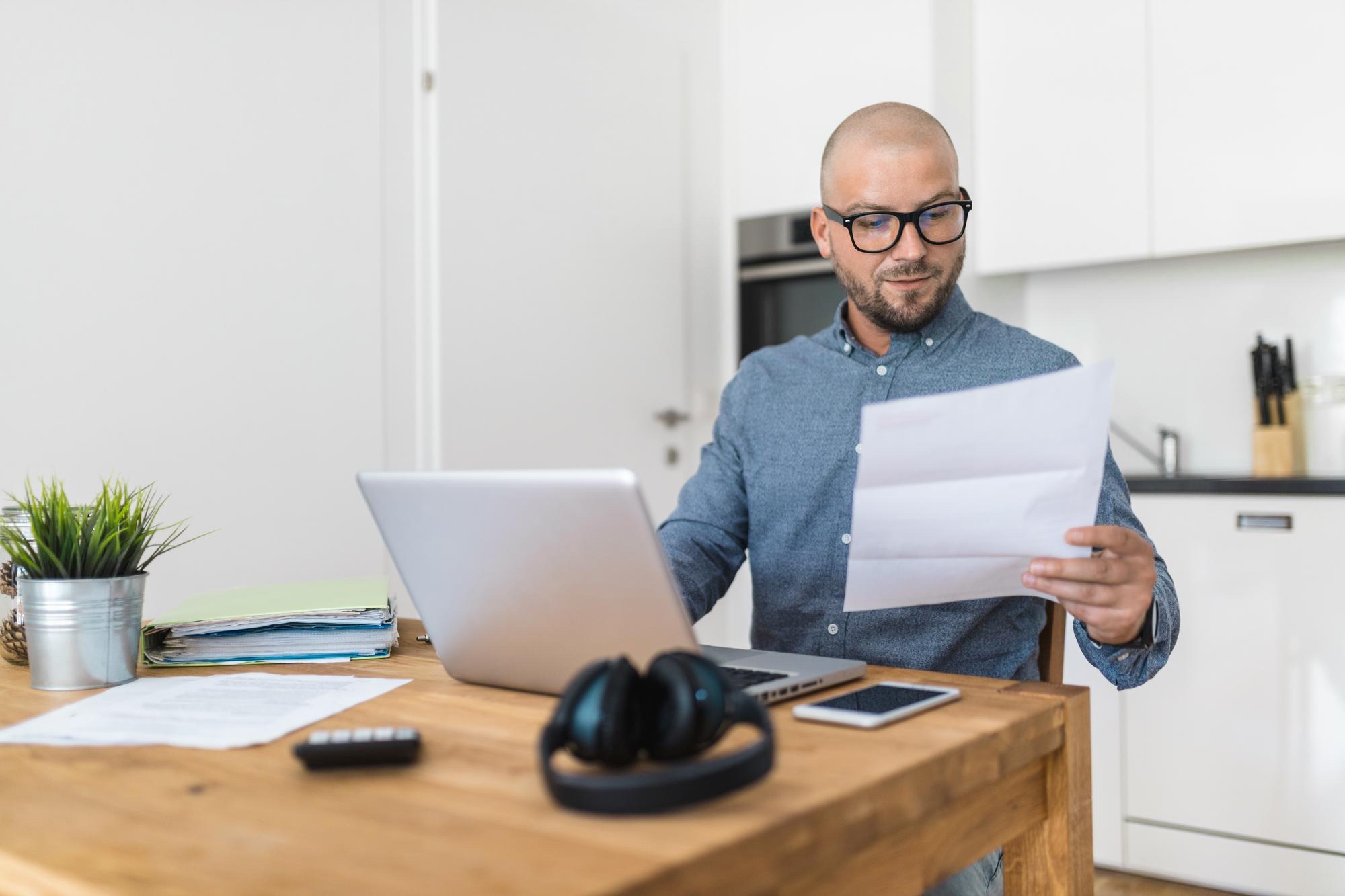 A man sits at a wooden table in a modern kitchen, looking at a document while working on a laptop. Headphones, a phone, paperwork, and a plant are also on the table.